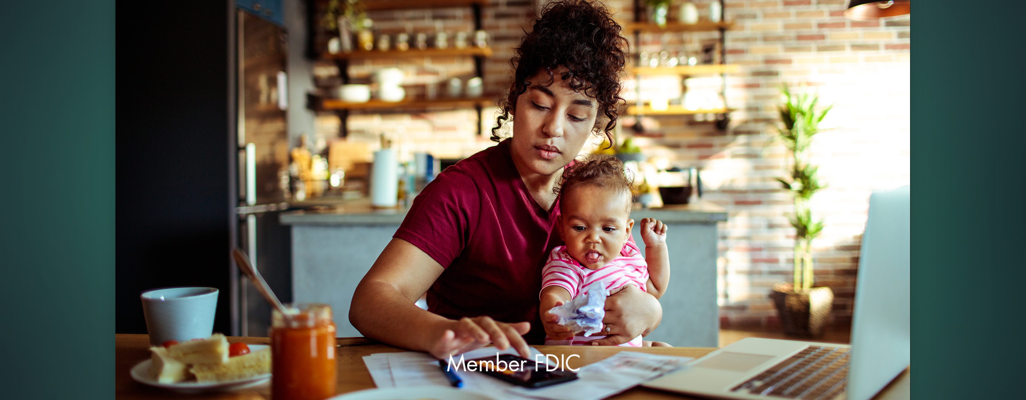 A mom holding her baby while going over her finances at the kitchen table. The lower part of the image reads: Member FDIC