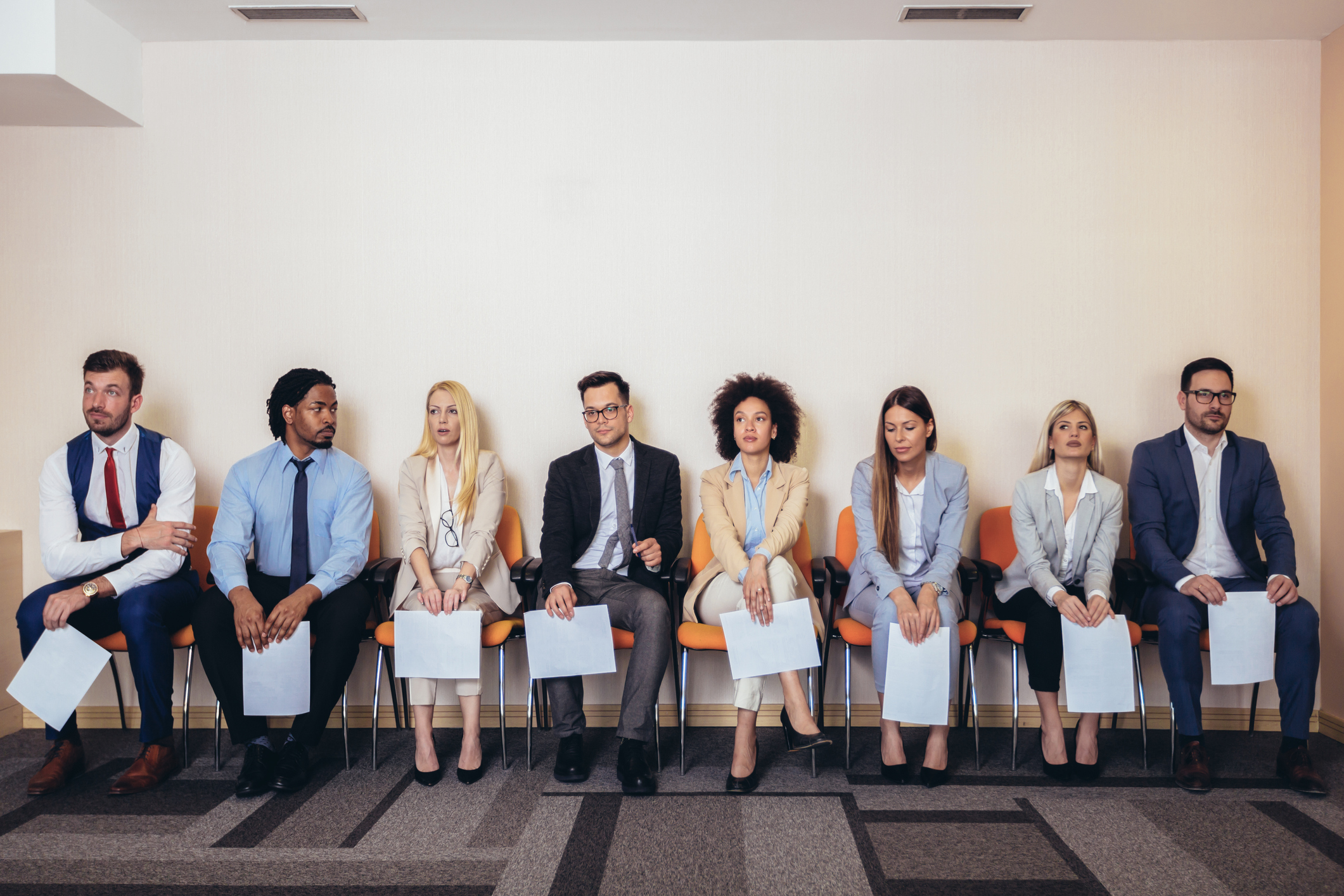 Eight candidates sitting in a row of chairs holding their resumes and waiting to be interviewed for a job
