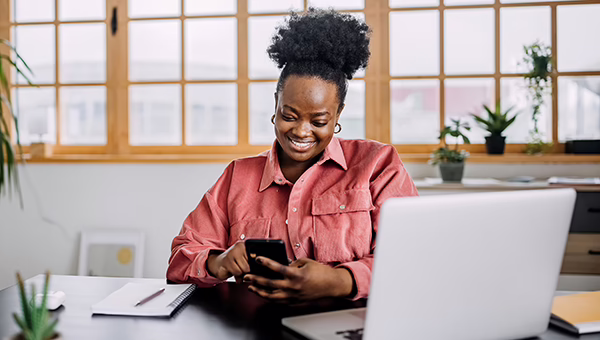 A woman sitting at a desk looking at her cell phone with a laptop on the desk in front of her