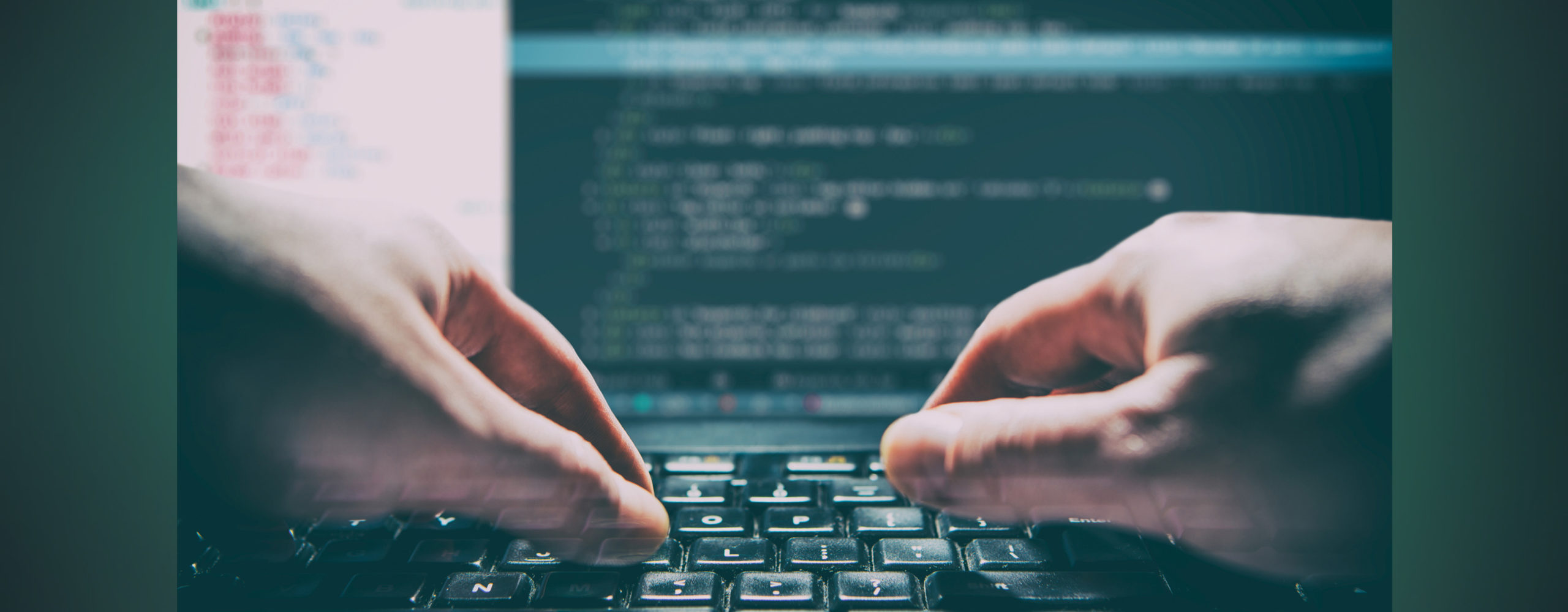 Close-up of hands typing on a keyboard with blurred code on a monitor in the background.