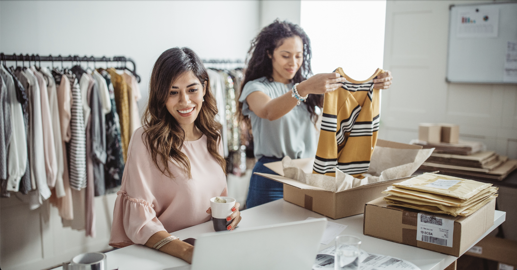 Two women working in a small boutique business. One is packing orders and the other is looking at her laptop.