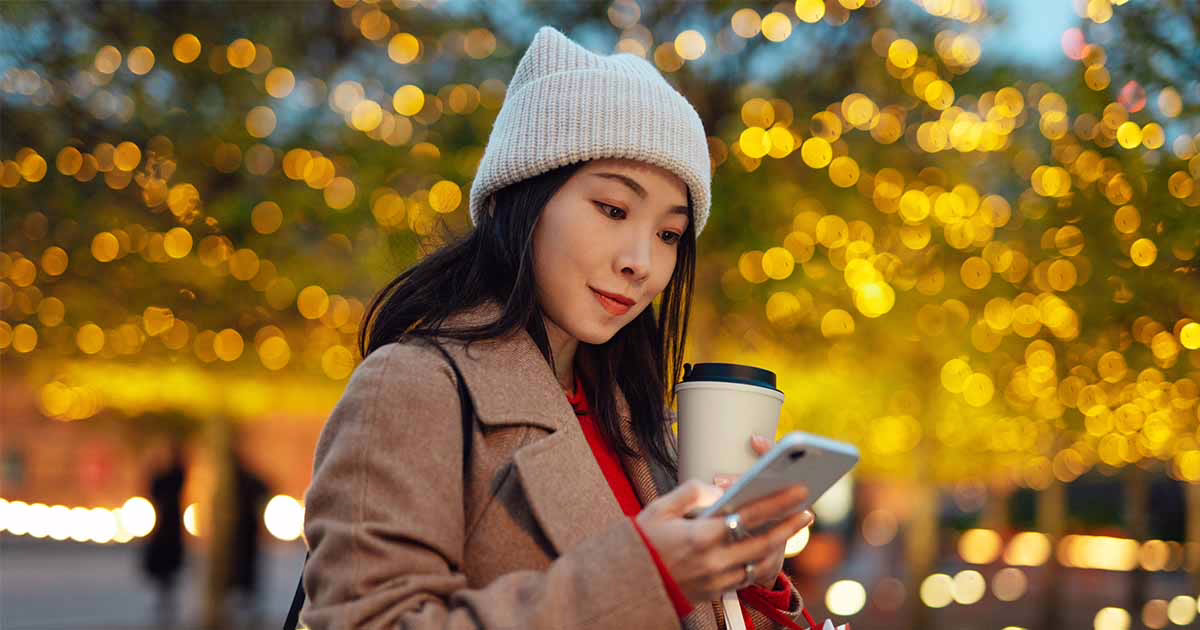 A woman wearing a winter coat looking at her phone with Christmas lights in the background